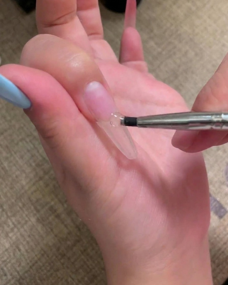 A brush applying a coat of flesh-pink gel polish onto a prepped nail.