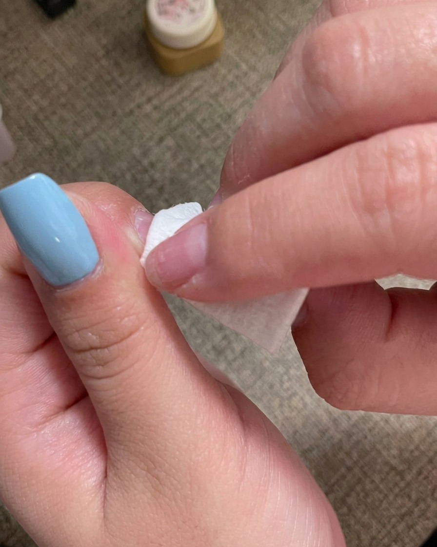 A hand using a lint-free wipe to clean the surface of a manicured nail.