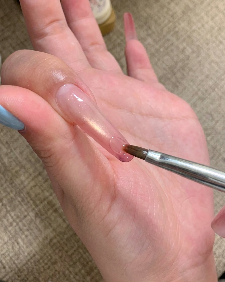 A nail art brush applying a drop of clear adhesive gel to the tip of a long nail.