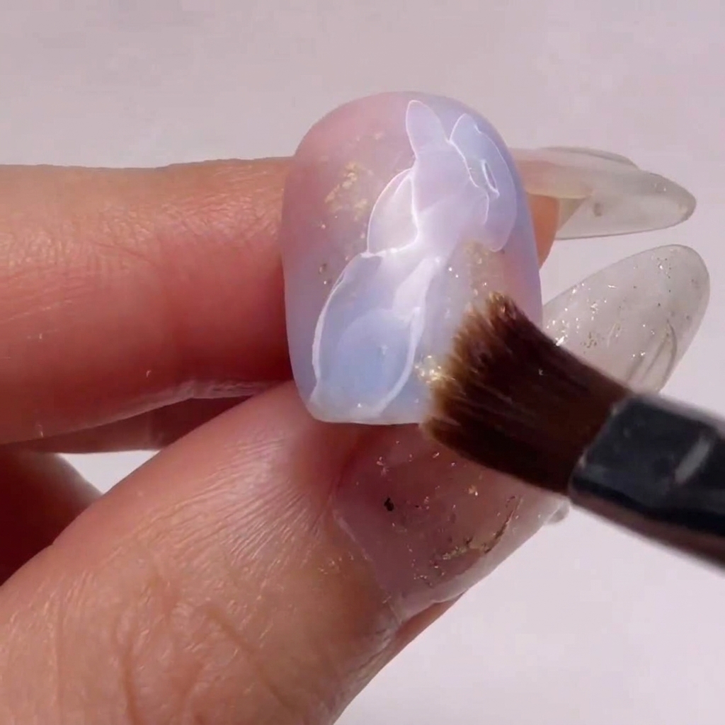 A close-up of a small nail brush using alcohol to gently blur and soften one edge of white gel nail art over a pink and blue gradient base.