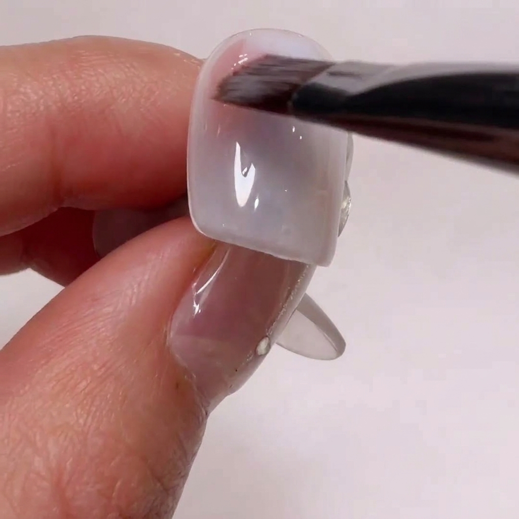 A nail art brush applying a soft nude pink gel to the side corners of a nail tip.