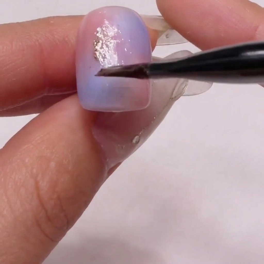 A brush carefully placing fine gold glitter particles onto the soft pink and blue gradient nail surface.