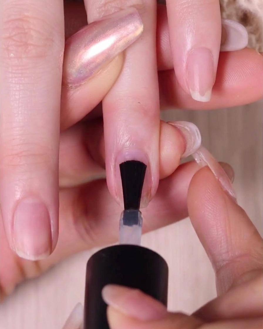 A close-up of a nail polish brush applying a clear base coat onto a bare natural fingernail.