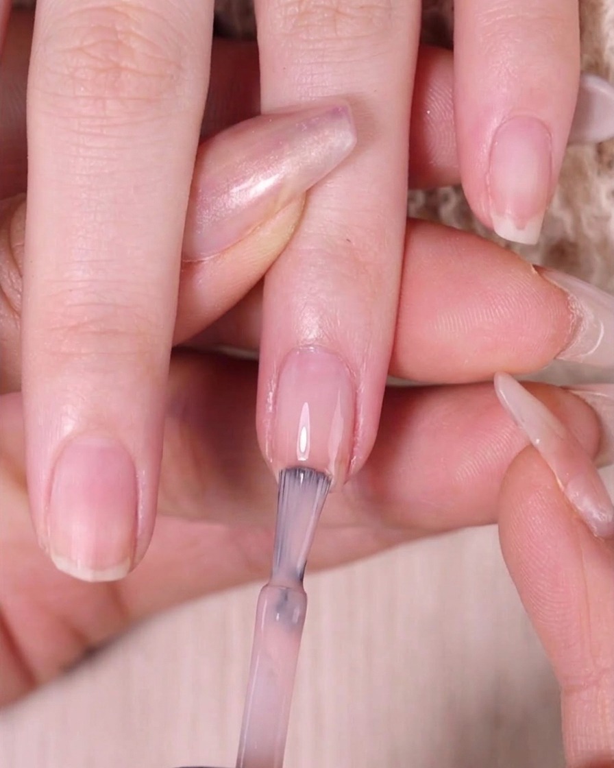 A nail brush applying sheer nude pink polish strictly to the top third of a fingernail.
