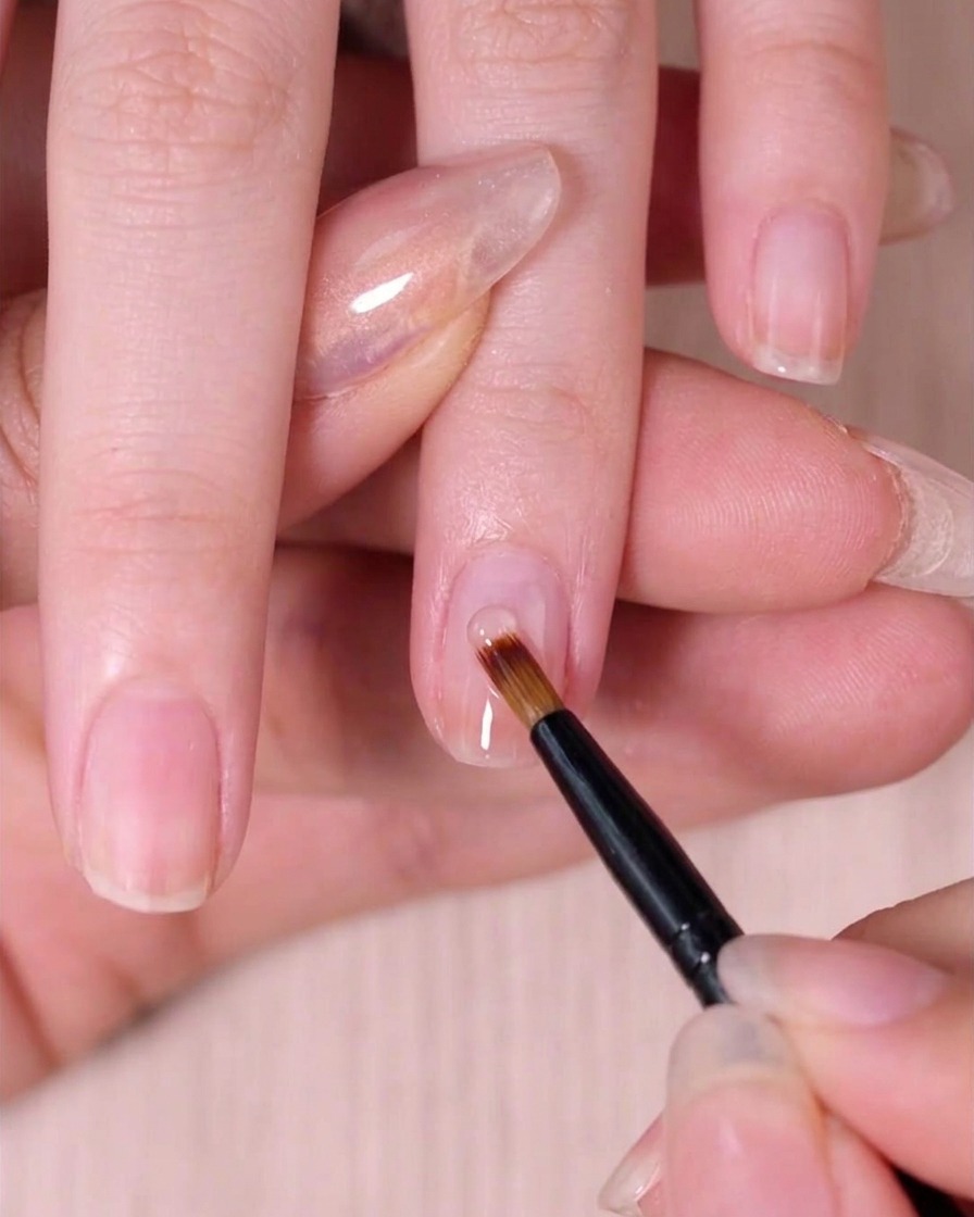 A specialized sweeping nail brush lightly blending wet pink polish down the nail to create an ombre effect.