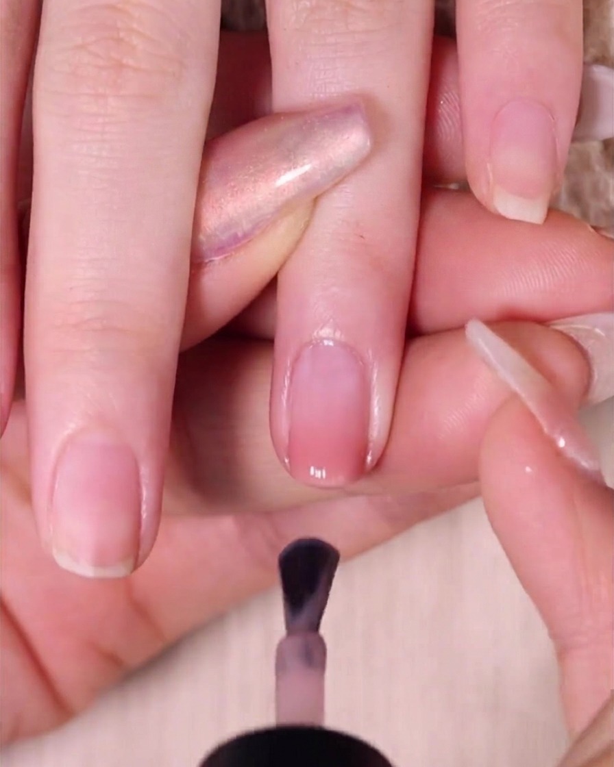 A nail brush applying a smooth coat of sheer nude pink polish over an ombre nail, leaving a small gap near the cuticle.