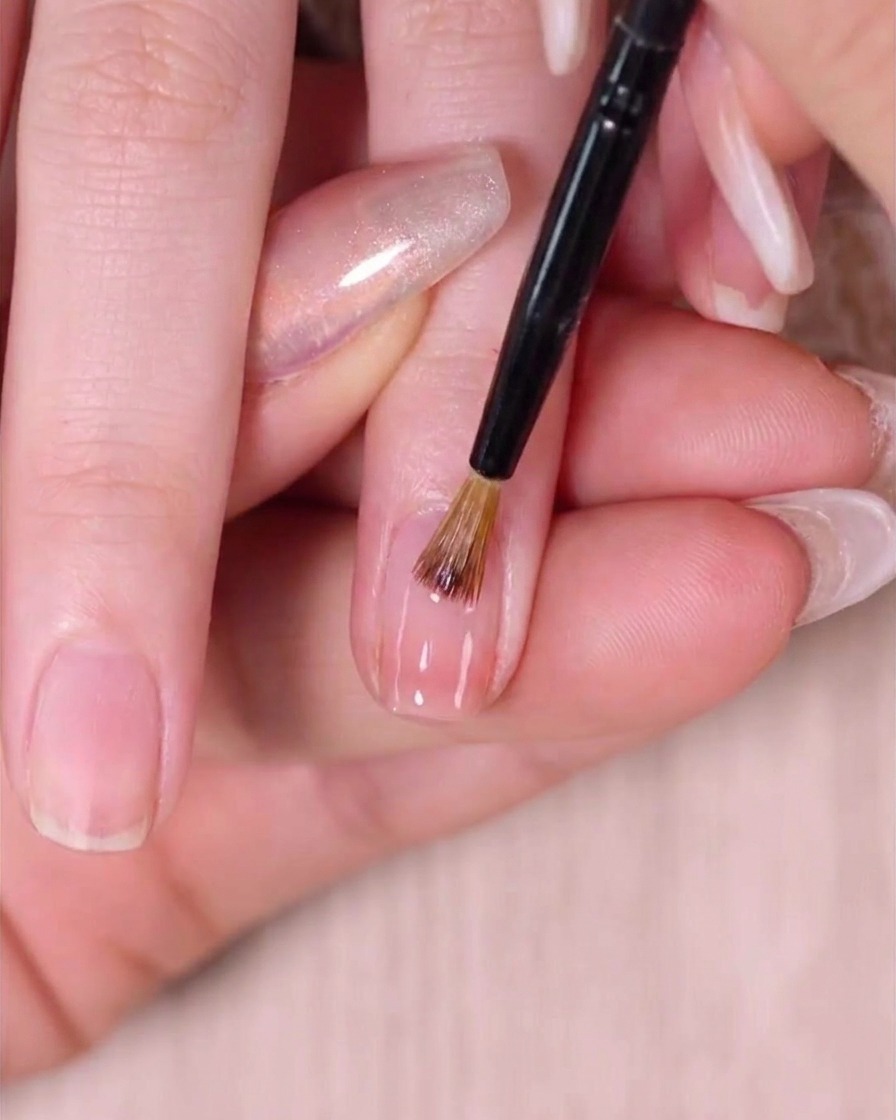 A fine-tipped nail art sweeping brush delicately feathering wet pink polish near the cuticle to create a seamless blend.