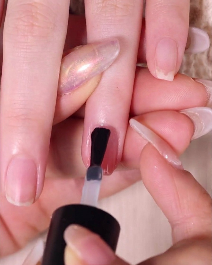 A brush applying a thick, glossy layer of clear top coat over a finished nude pink ombre fingernail.