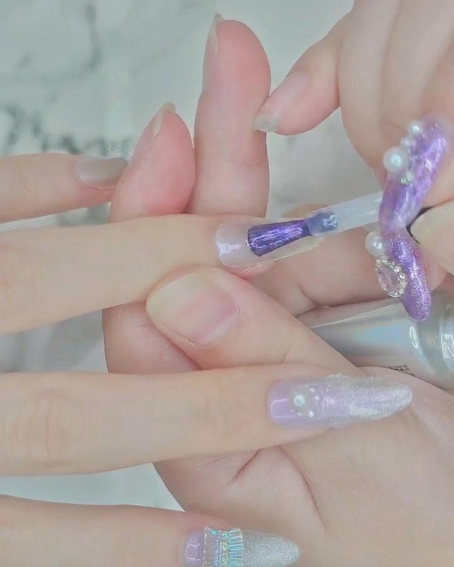 A close-up view of a brush applying sheer purple gel polish to the center of a clear nail extension.