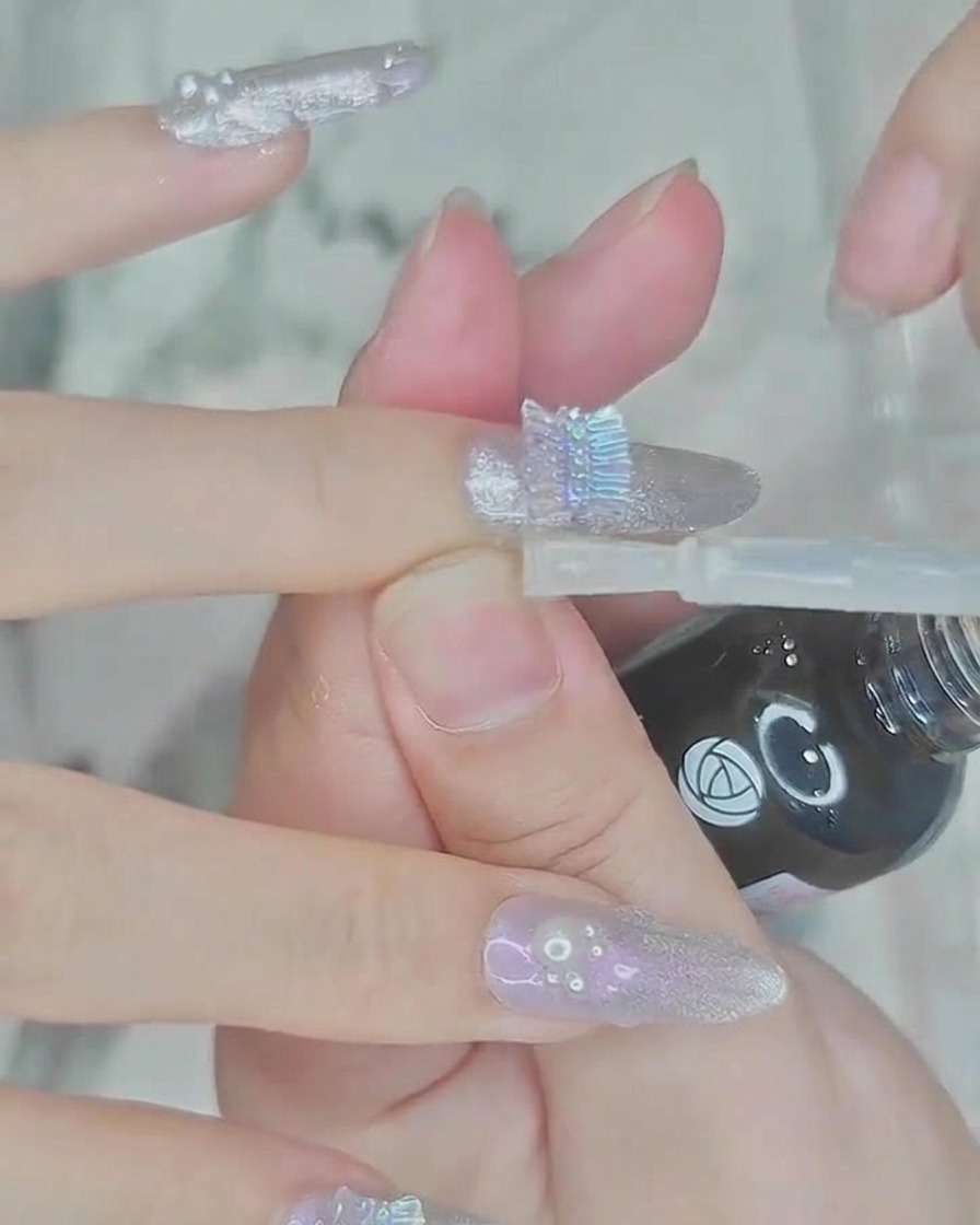 A brush applying a layer of clear glossy top coat over the nail and pearl embellishments.