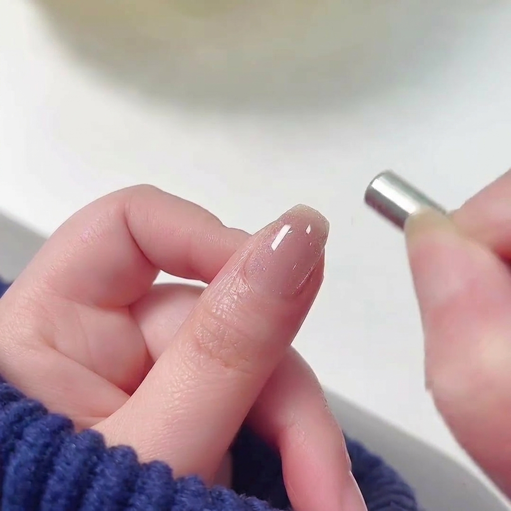 A small cylindrical magnet being held near the cuticle of a fingernail coated in wet, shimmering pink cat-eye gel polish.