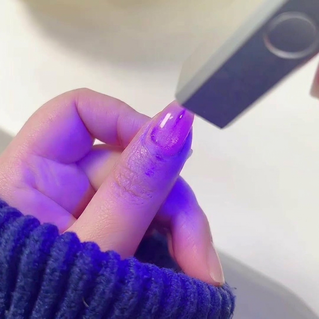 A finger with wet, shimmering purple gel polish positioned inside a glowing UV/LED nail lamp.