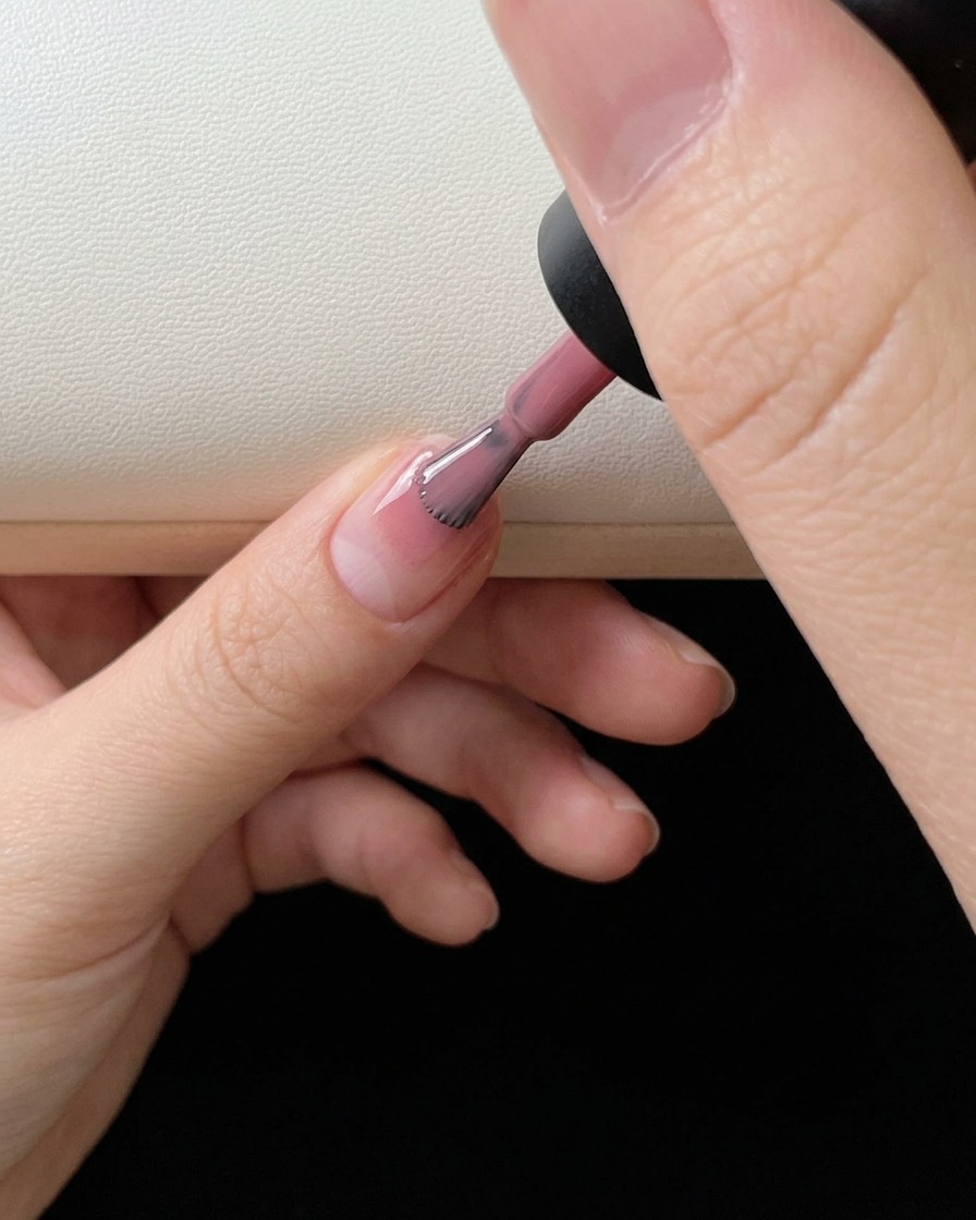 A close-up of a nail polish brush applying pink color gel to the tip of a natural fingernail.
