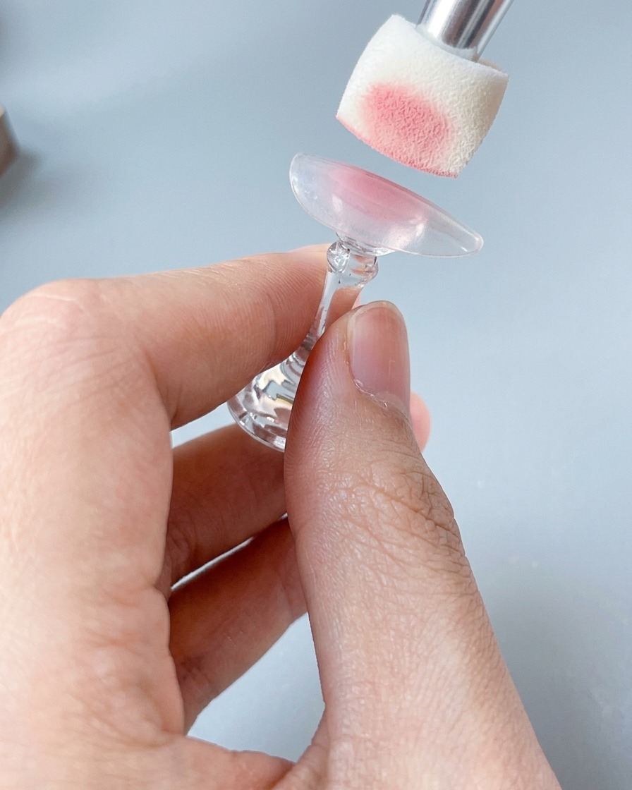 A close-up of a sponge tool applying a soft blush pink color to a nail tip held on a stand.