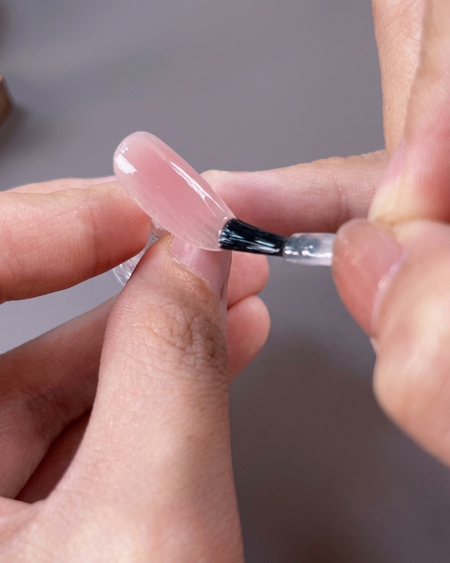 A nail brush applying a sheer, light pinkish-nude gel polish over the entire surface of the nail.