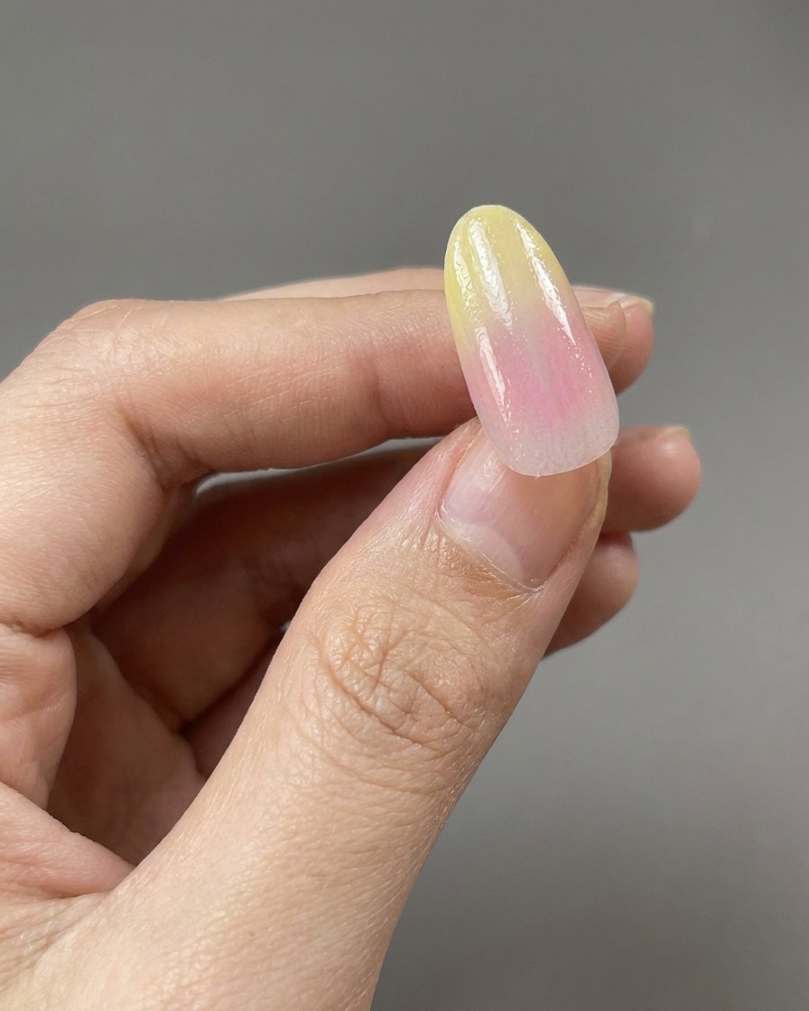 A brush applying a clear, glossy base coat over a yellow and pink gradient nail design.