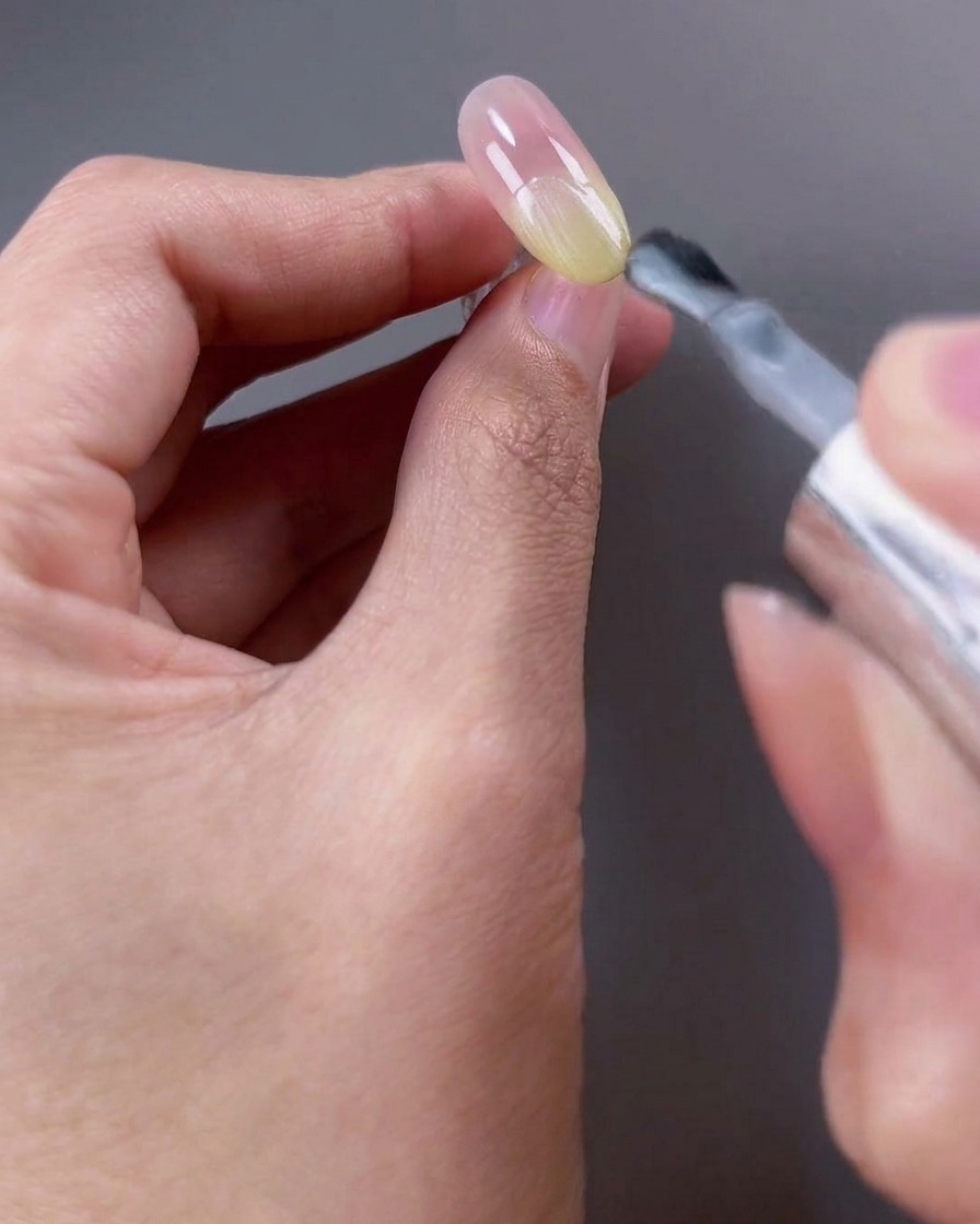 A nail brush applying a sheer white shimmer gel polish to the top half of the nail surface.