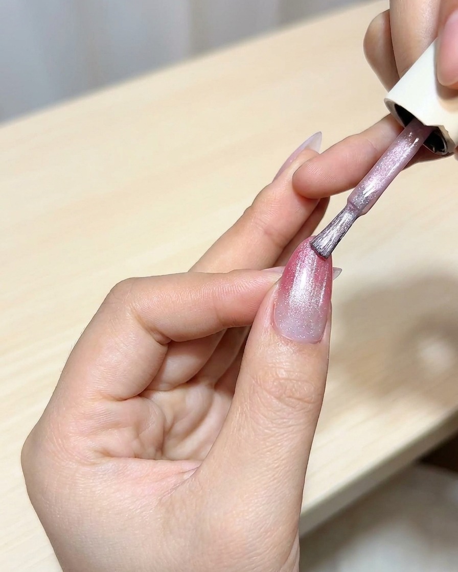 A brush applying a layer of cat-eye gel polish over a magenta ombre base.