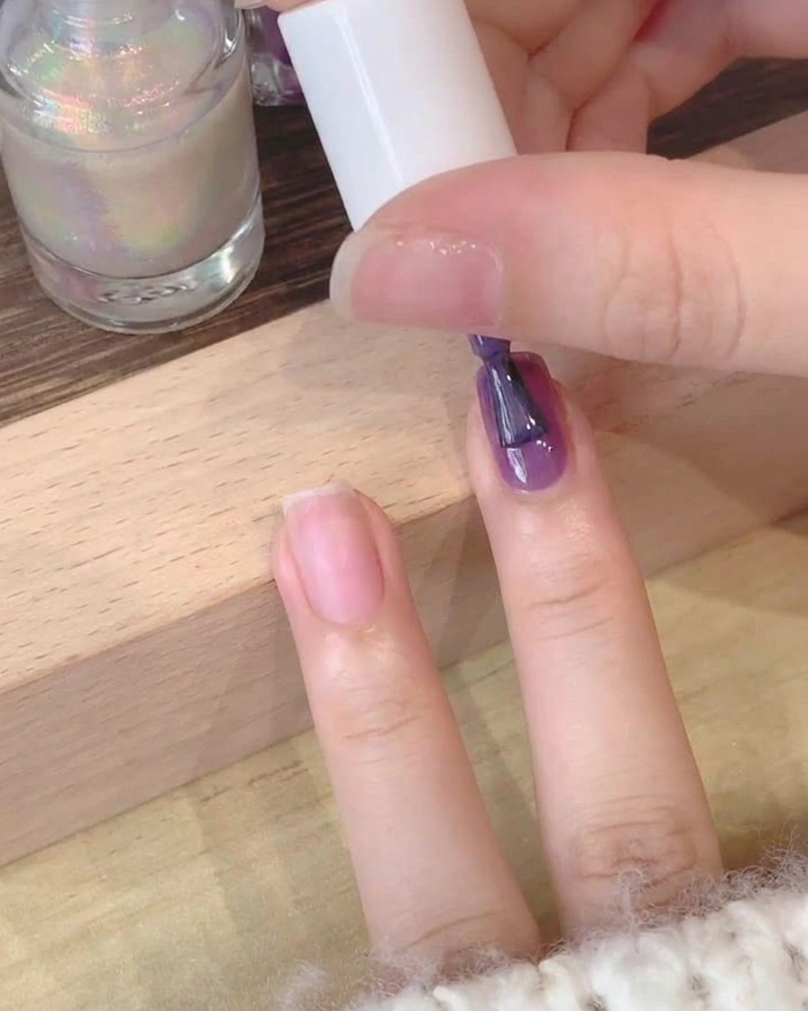 A close-up view of a brush applying deep purple gel polish to a natural fingernail.