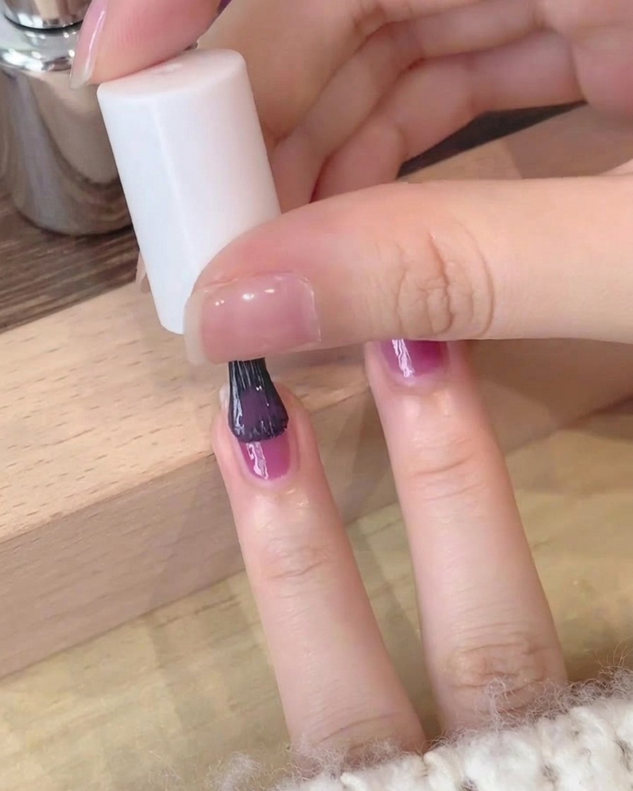 Close-up of a second layer of deep purple gel polish being carefully applied to a fingernail.