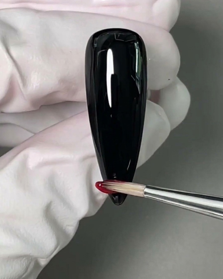 A small nail art brush applying a vibrant red gel polish to the tip of a black-painted nail.