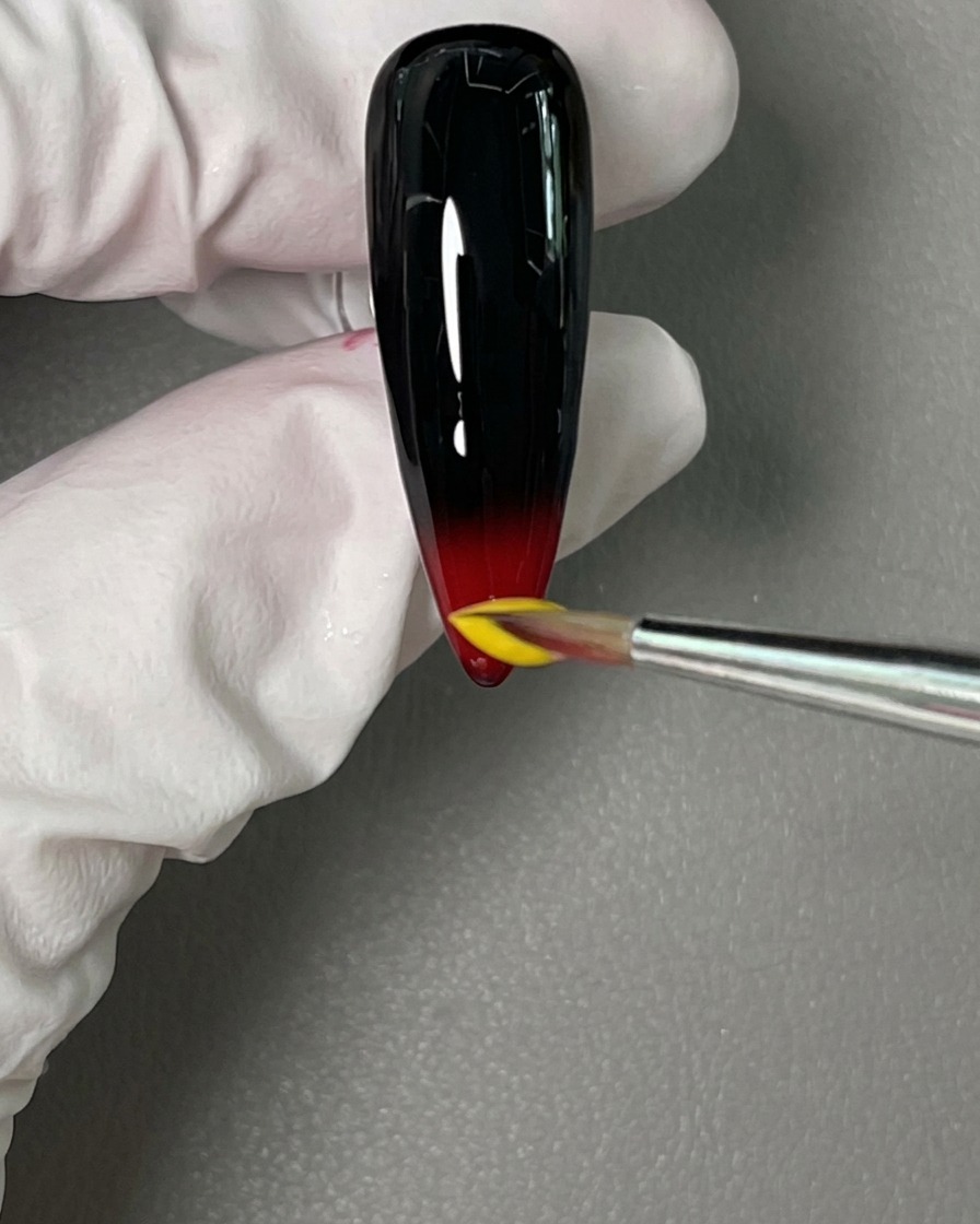 A detail brush placing a bead of bright yellow gel polish directly above the wet red gel on the nail tip.
