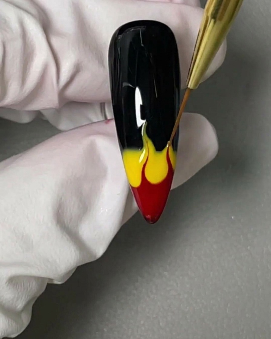A fine-tipped liner brush pulling wet red and yellow gel polish upwards on a black nail to form flame shapes.