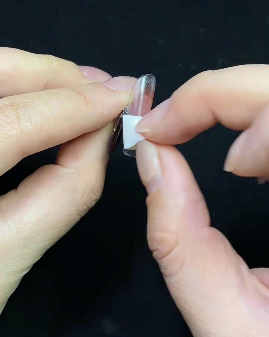 A close-up of a brush applying a nude-colored gel base coat to a clear plastic nail tip.