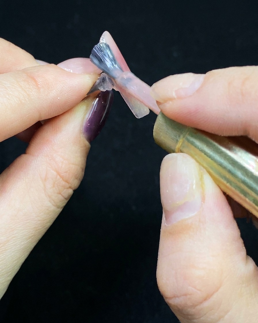 A brush applying a thin layer of nude gel polish over a clear nail tip.