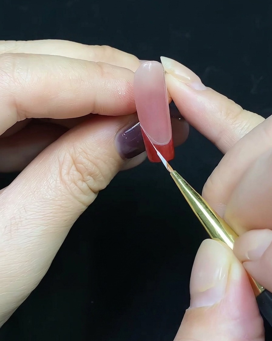 A fine liner brush drawing a thin white line along the edge of a red French tip.