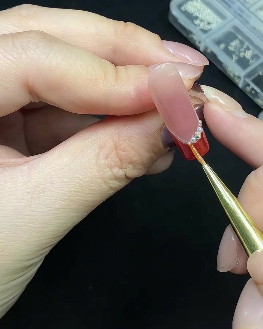 A precision tool placing small pearls along the base of a red French tip nail.