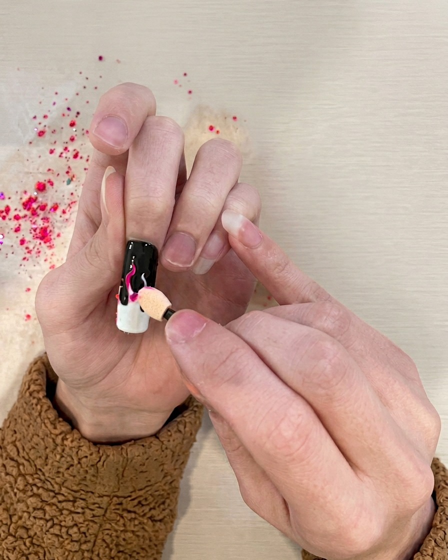 A sponge applicator dabbing vibrant pink pigment onto the white flame details of the nail.
