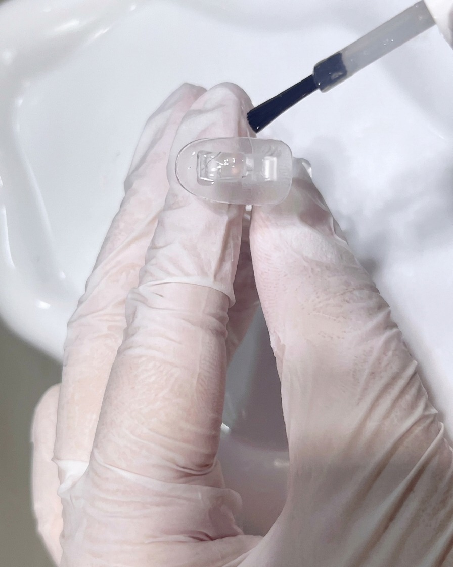 A close-up view of a brush applying a clear base coat onto a clear nail tip.