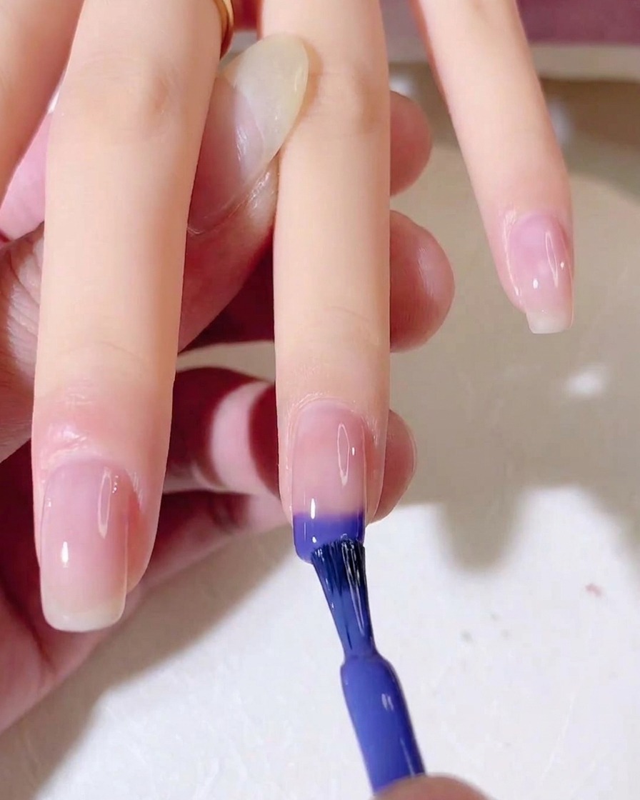 A nail brush applying a blue and purple gel gradient onto the tip of a natural fingernail.