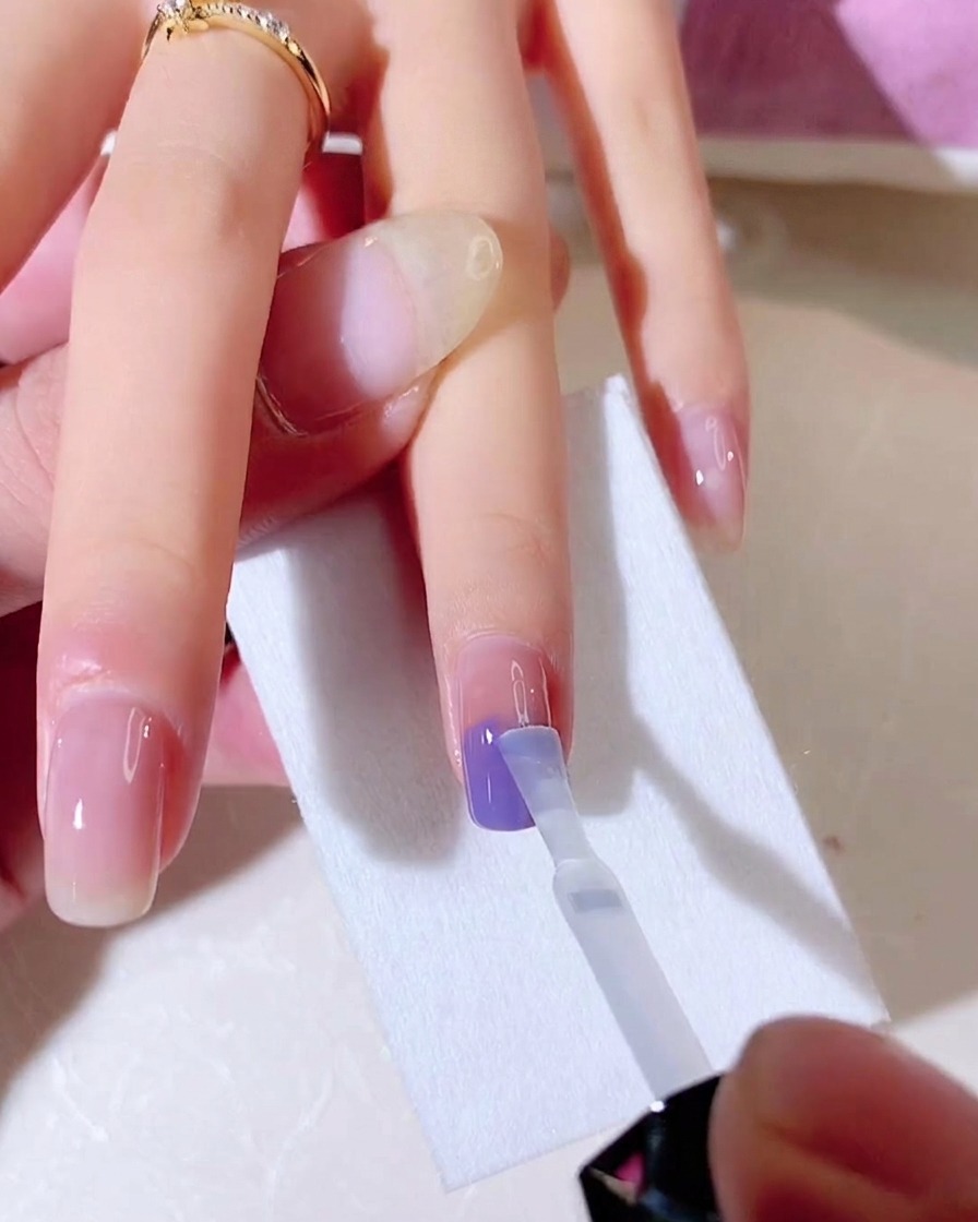 A clear gel base coat brush being used to blend blue and purple colors vertically on a nail.