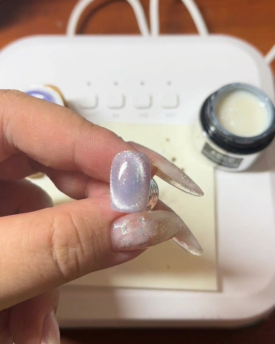 A close-up of a hand holding a magnetic wand over a purple cat-eye gel nail, showing the bright cat-eye streak forming.