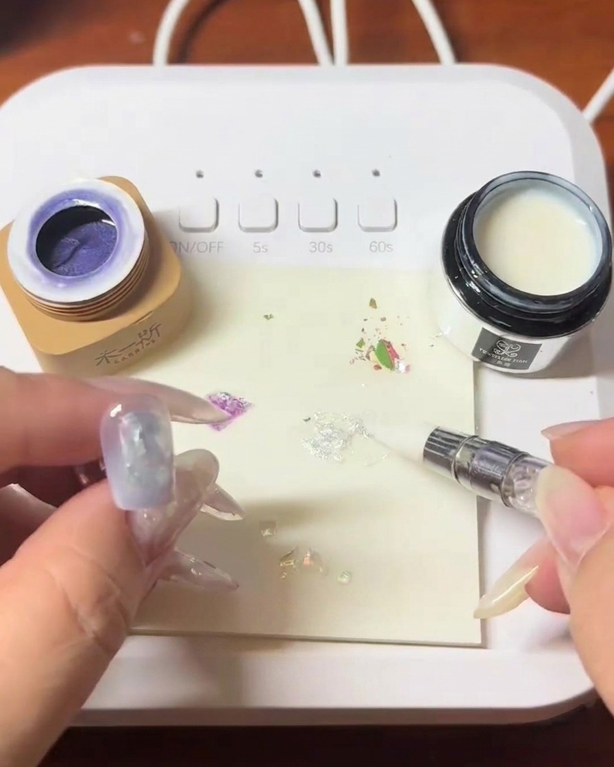 A brush applying a clear blooming gel over the shell-embellished nail.