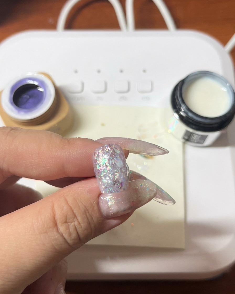 A brush applying purple gel polish over a nail already decorated with glitter and shell.