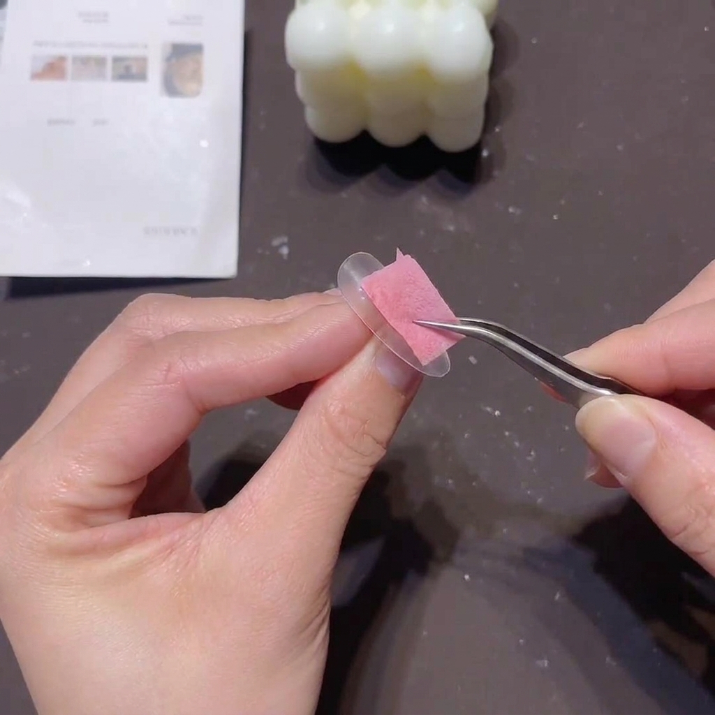 A person using tweezers to hold a small pink sponge, dabbing a light concealer gel onto the free edge of a clear nail tip.