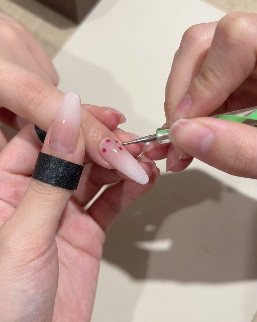 A close-up of a dotting tool applying precise, small red polka dots onto a soft pink gradient base nail.