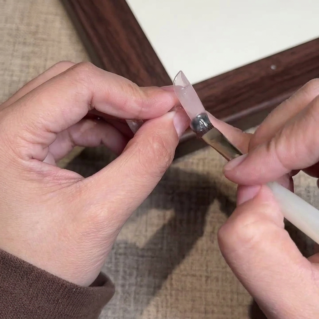 A close-up of a brush applying a sheer nude pink gel polish onto a natural fingernail.