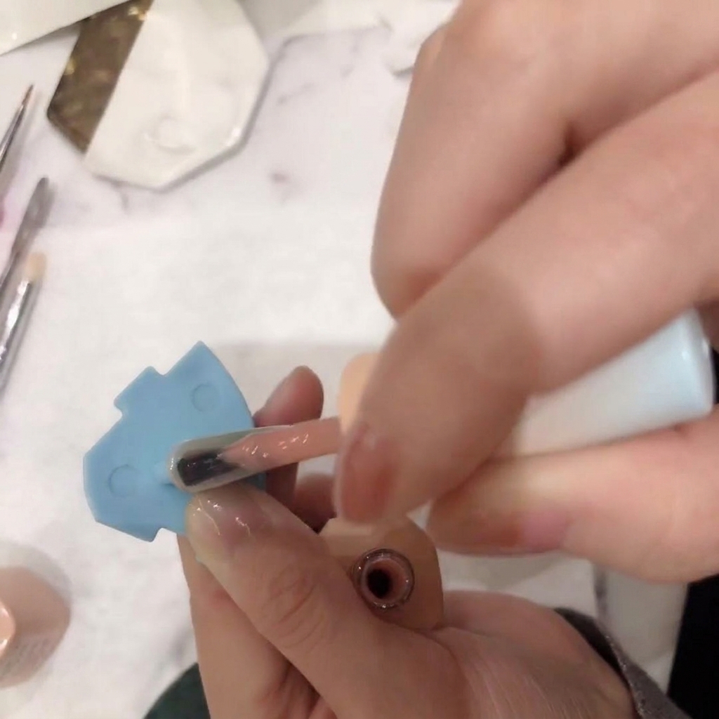 A close-up of a brush applying a sheer, nude pink gel polish onto a nail.