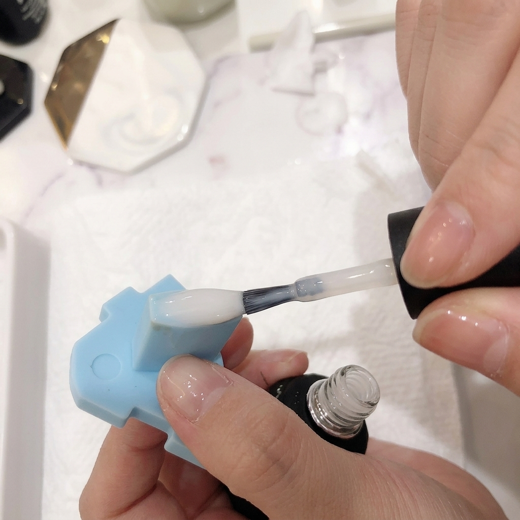 A close-up of a brush applying a sheer layer of milky white polish over a pink and white ombre nail.
