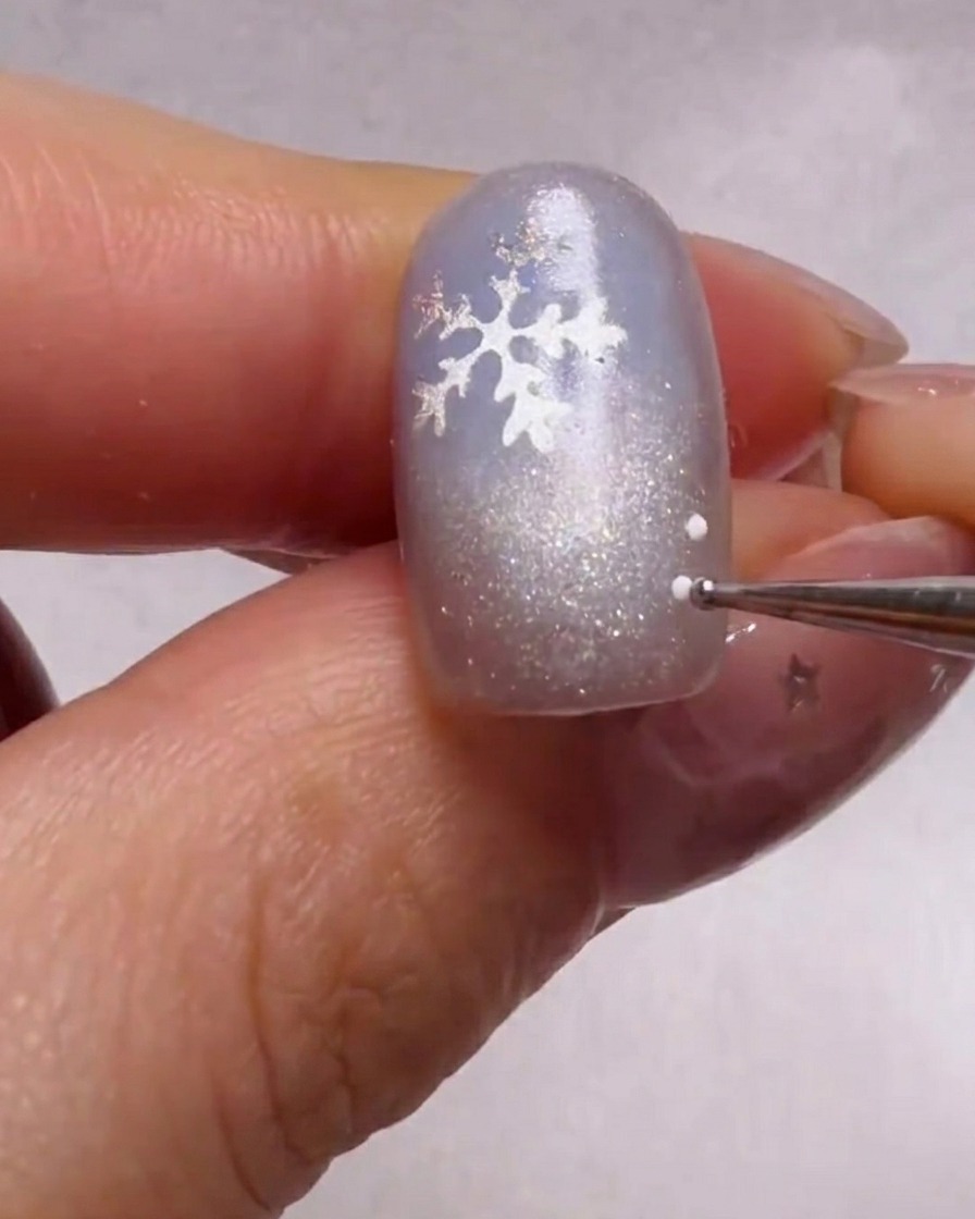 A close-up view of a dotting tool applying tiny white and light blue dots around a silver snowflake sticker on a shimmery blue nail.