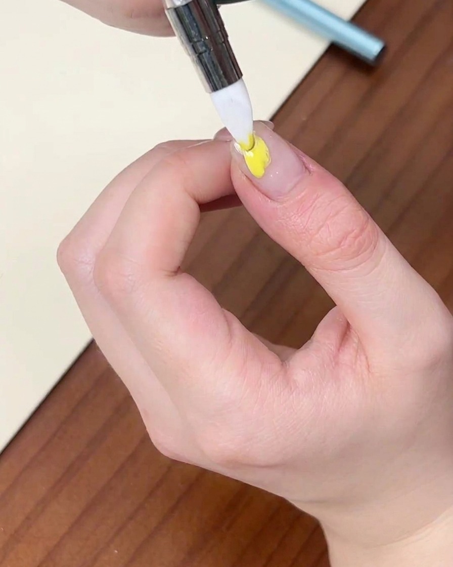 A fine liner brush applying a dot of yellow gel polish onto a natural fingernail.