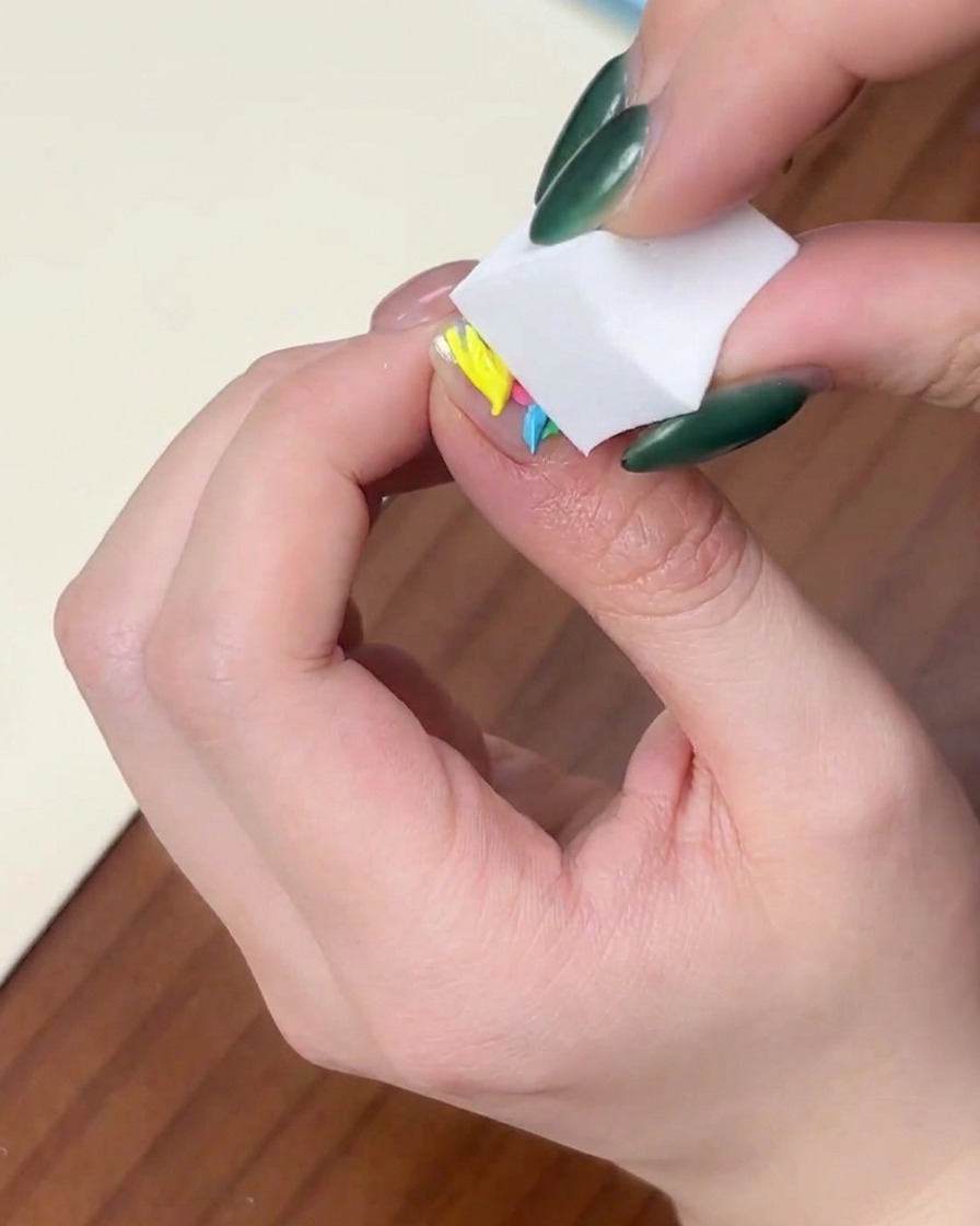 A close-up of a hand pressing a white makeup sponge vertically onto a fingernail with dots of colorful gel polish.