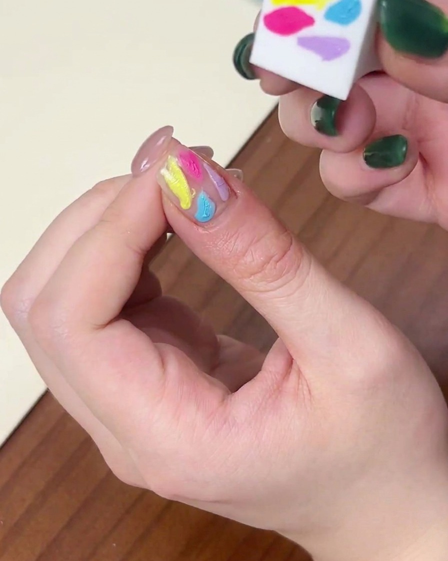 A close-up view showing the side of a sponge being used to blend multicolored gel polish dots on a fingernail.