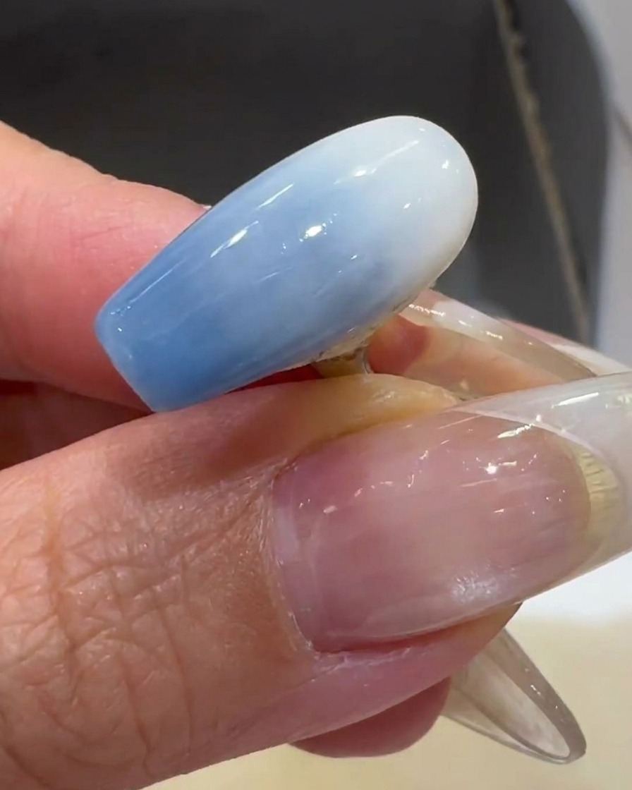 A brush applying a thin layer of milky white gel polish to a clear nail tip.