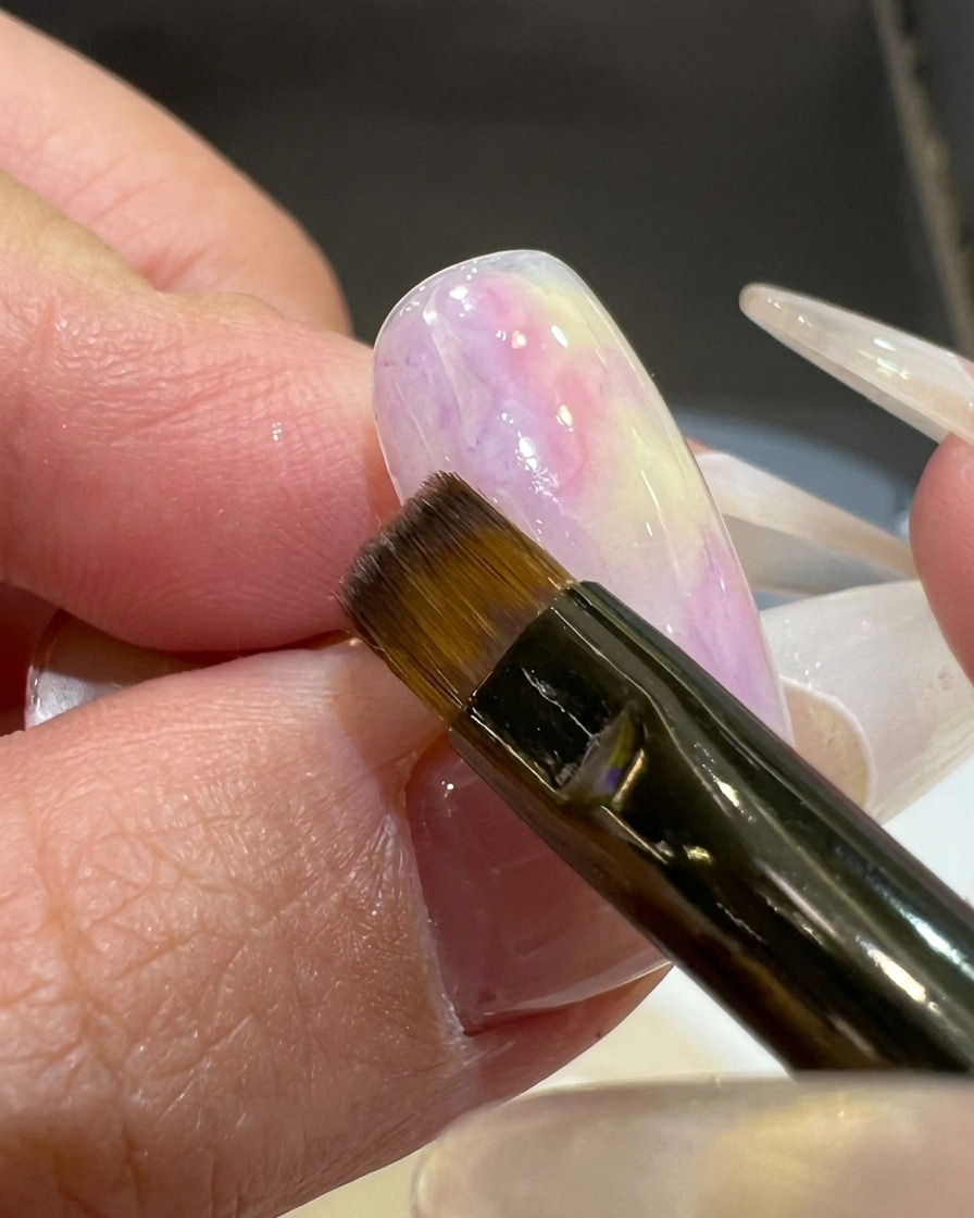 A flat brush applying a sheer pinkish-purple gel color to the corner of a white-based nail tip.