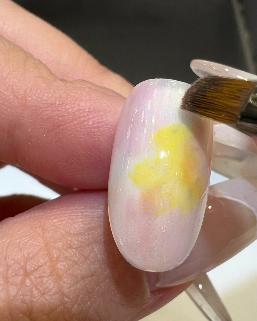A flat brush smoothing out the gradient between pink and yellow gel polish on a nail tip.